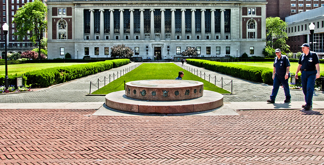 Two stacked concrete circle on a brick walkway. In the distance, a three separate lawns sit in front of a grand columned building.