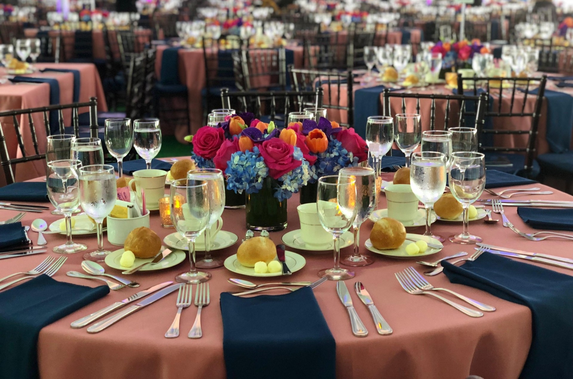 A banquet round is dressed with a pink tablecloth and navy blue napkins. Dinner rolls are placed at every place setting. At the center of the table is a fresh floral arrangement.