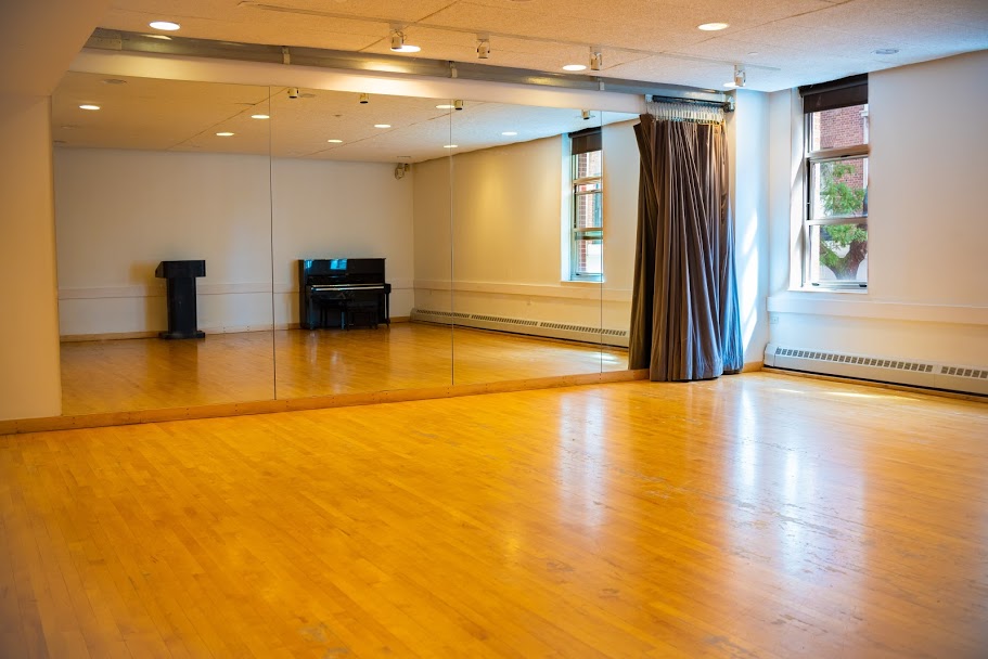 A room with wood floors and a wall of mirrors. In the mirror reflection there is a podium and piano. 