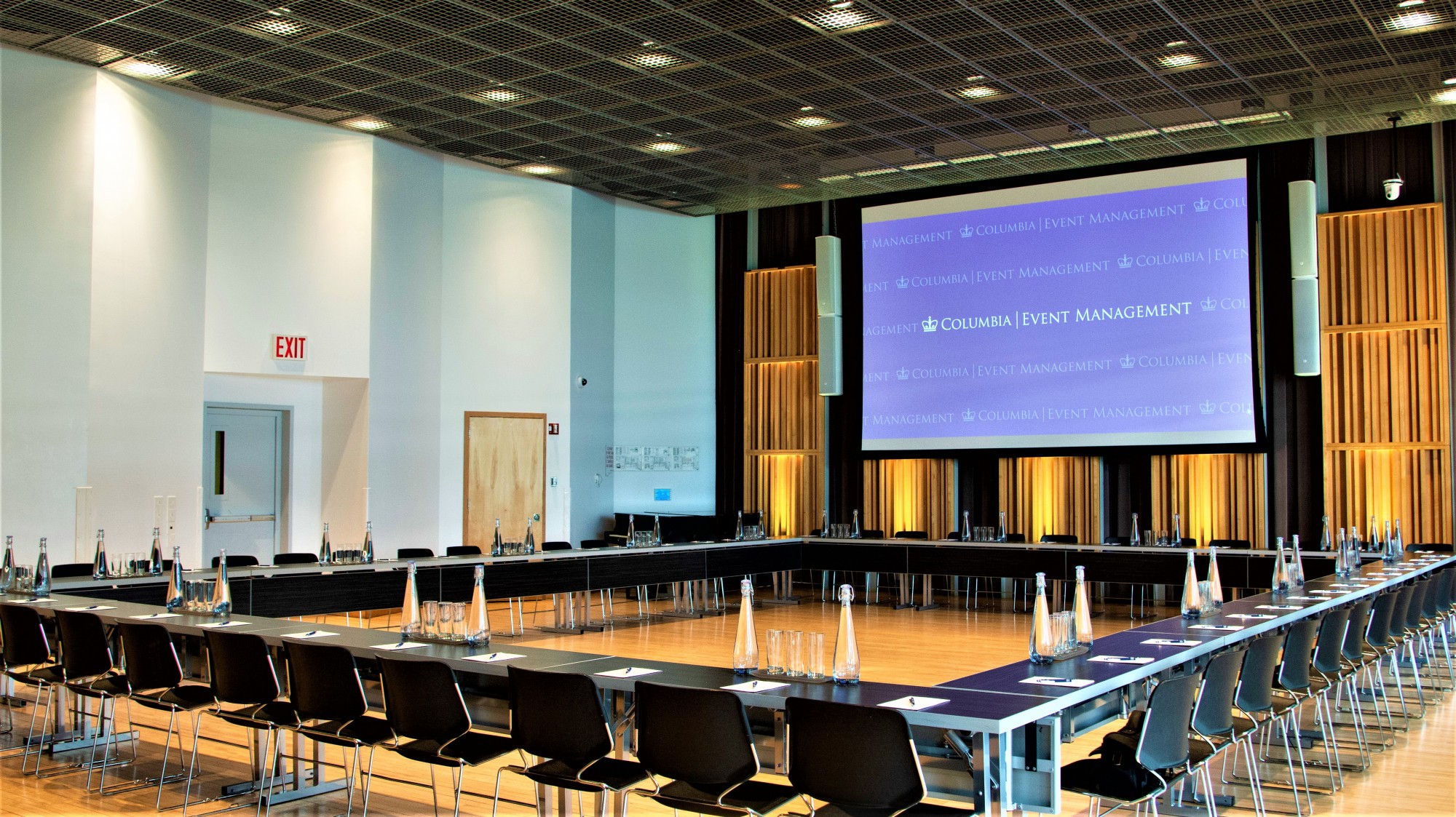 A large room has long tables arranged into a square shape. The tables are set with water glasses and pitchers. On one wall, a projector screen displays text, "Columbia University"