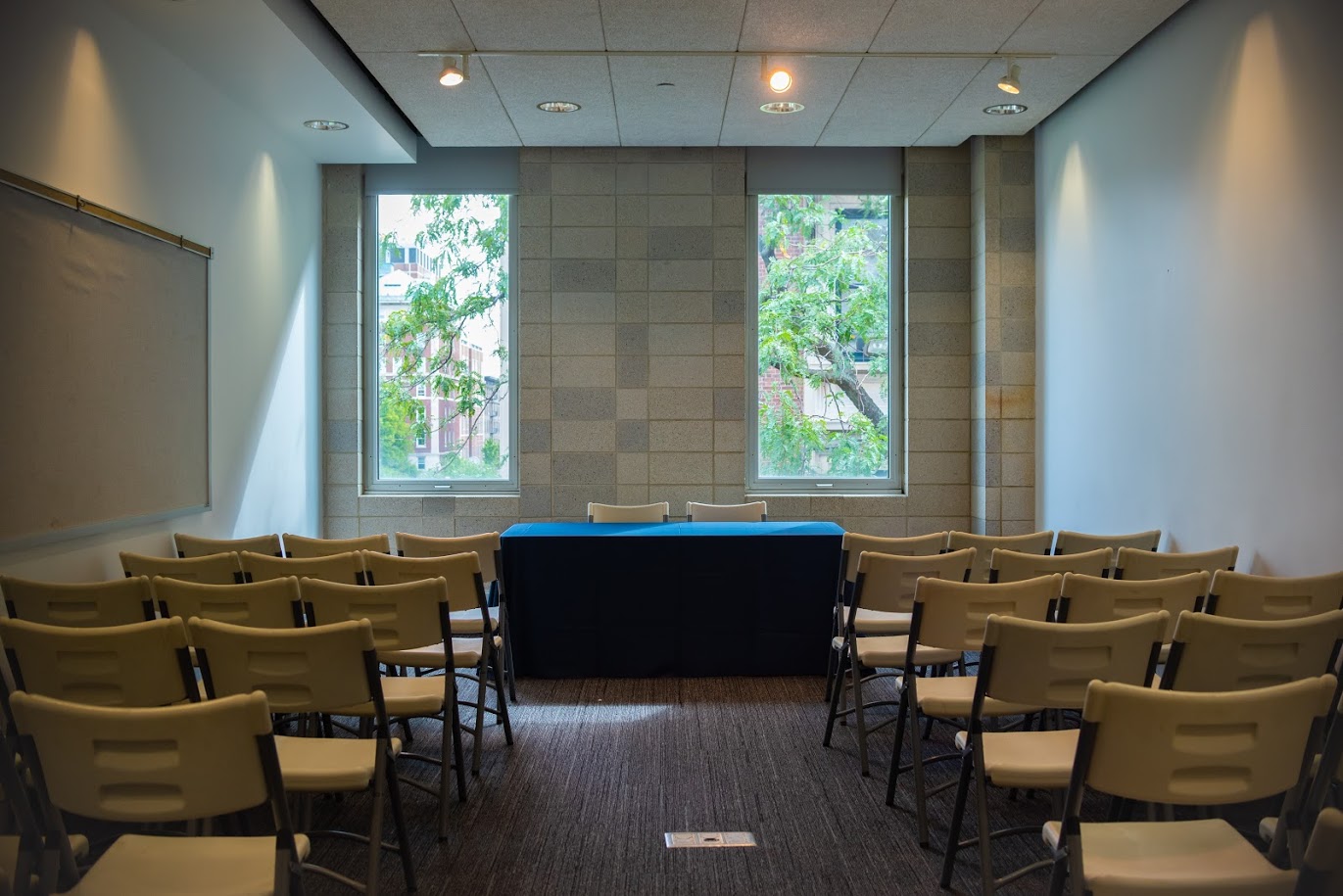 A grey-carpeted room with rows of white folding chairs facing a table covered with a blue table cloth. Two windows allow natural light into the room.