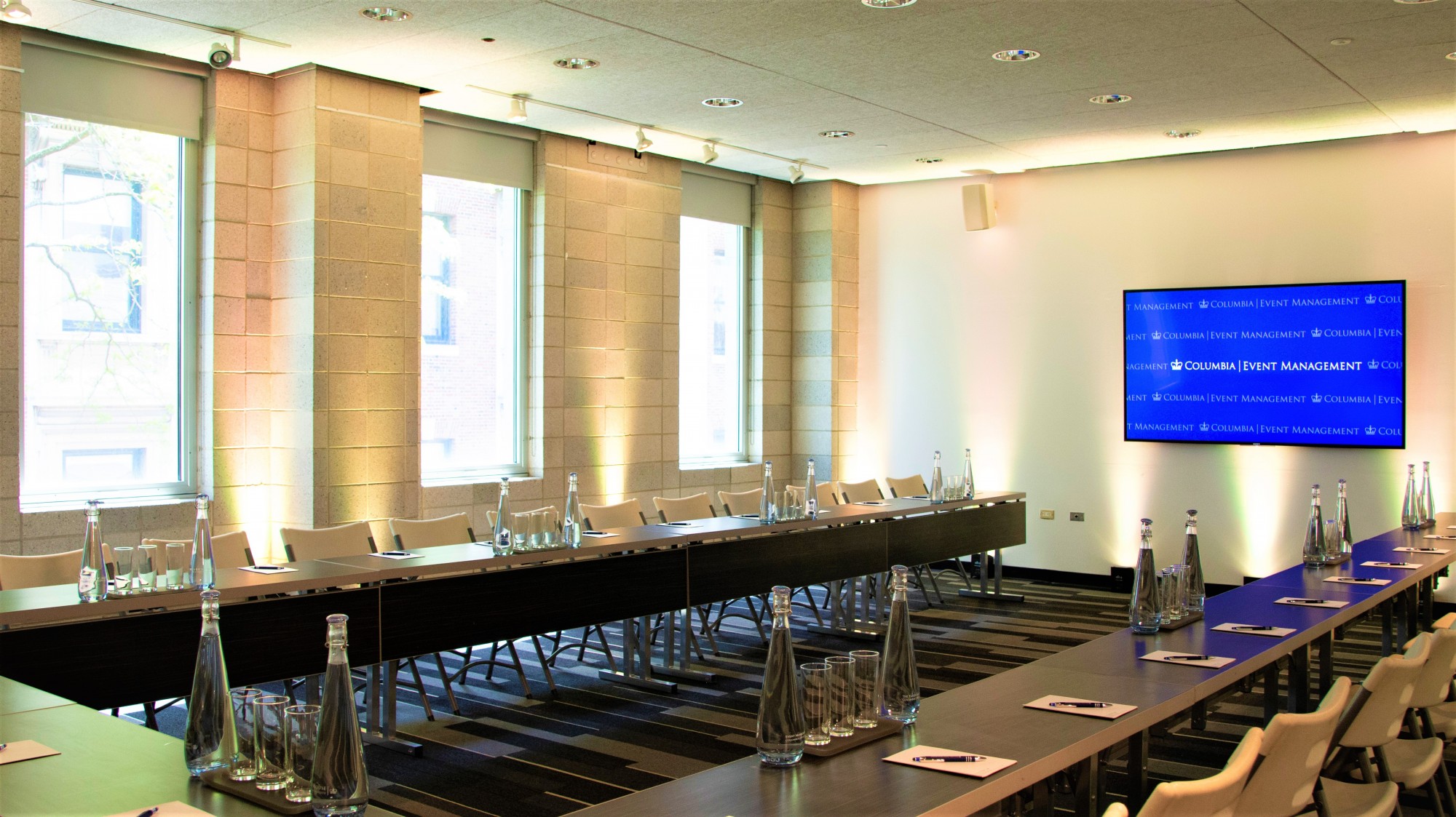 Long tables are arranged into a U-shape and set with water glasses and pitchers. . A TV screen is mounted on the wall facing the tables.