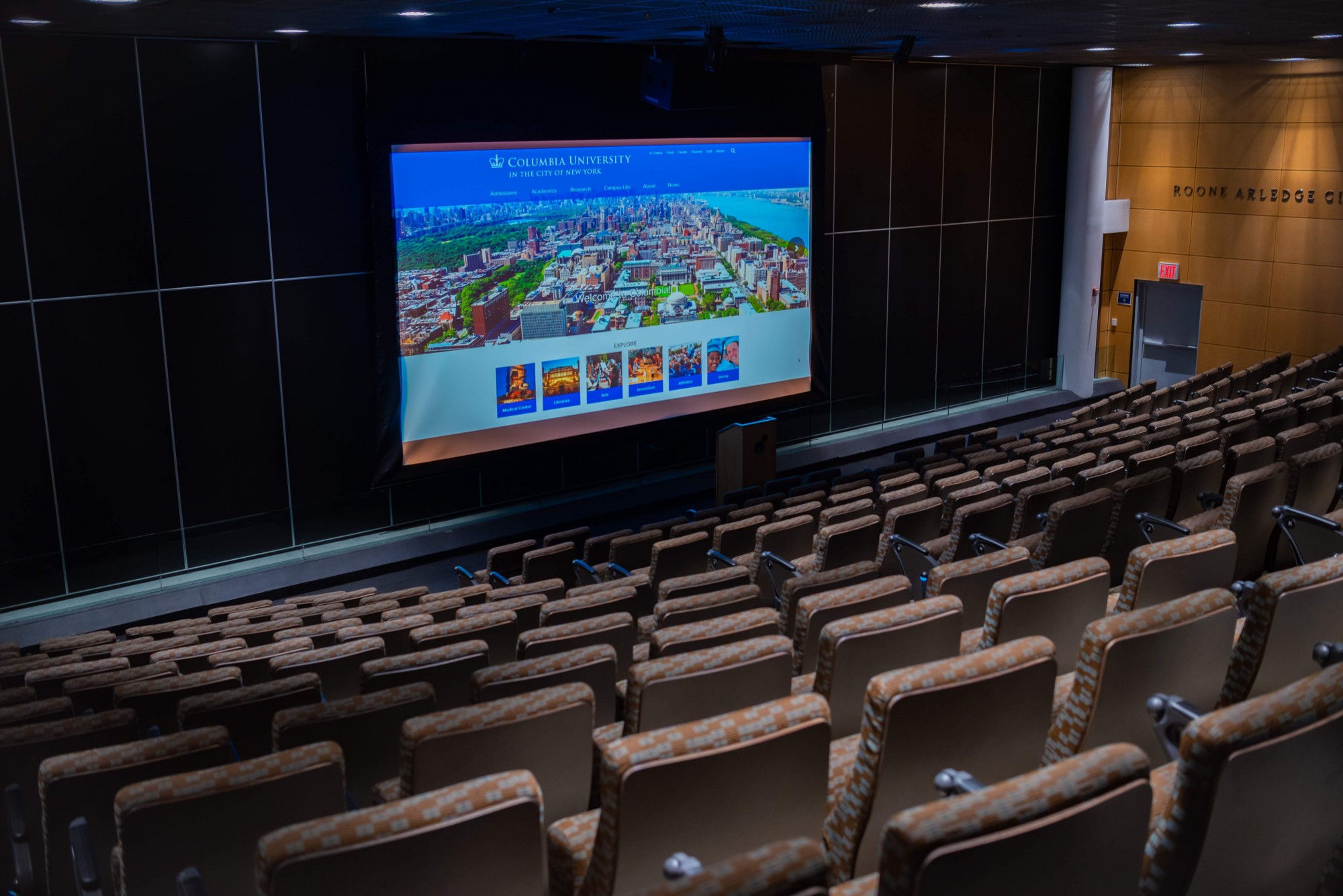 The picture shows an auditorium with theater-style seating. Rows of blue chairs face a large projector screen that shows the Columbia lion. 