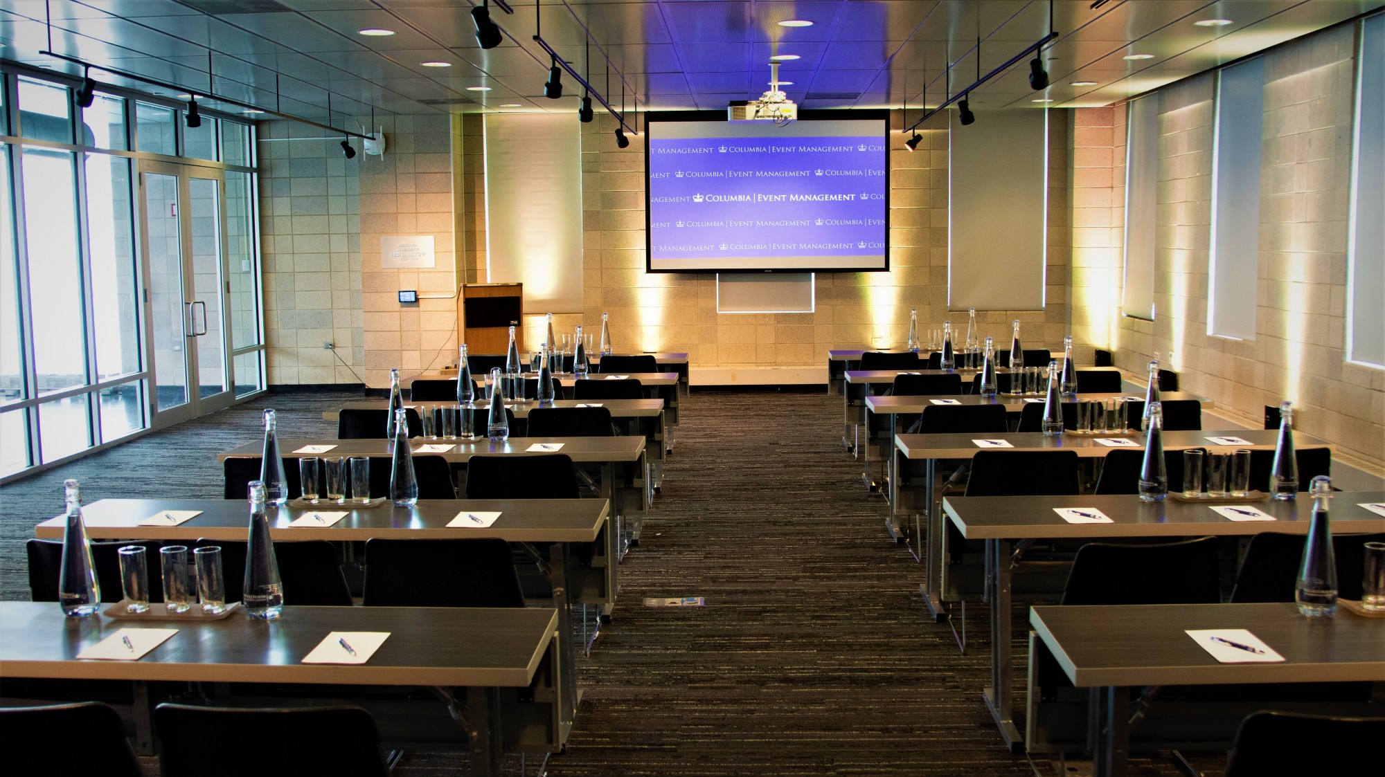 Rows of black tables set with two chairs, water glasses, and water pitchers face a projector screen.