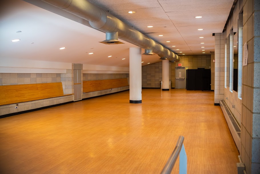 A large, open room with wooden floors, a sloped ceiling, and columns. Built-in wooden benches line the back wall.