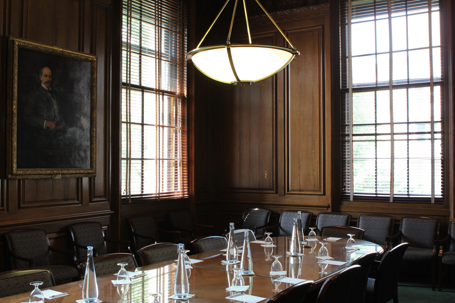 A long conference table set with glasses and water bottles sits in the middle of a wood-paneled room. Chairs are lined around the table and against the walls.
