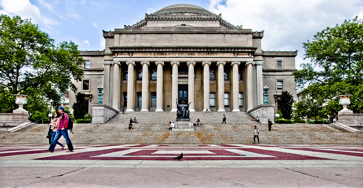 A grey and red brick patterned plaza sits in front of grand stairs and an ornate grey stone, columned building. People are walking across the plaza and sitting on the stairs.