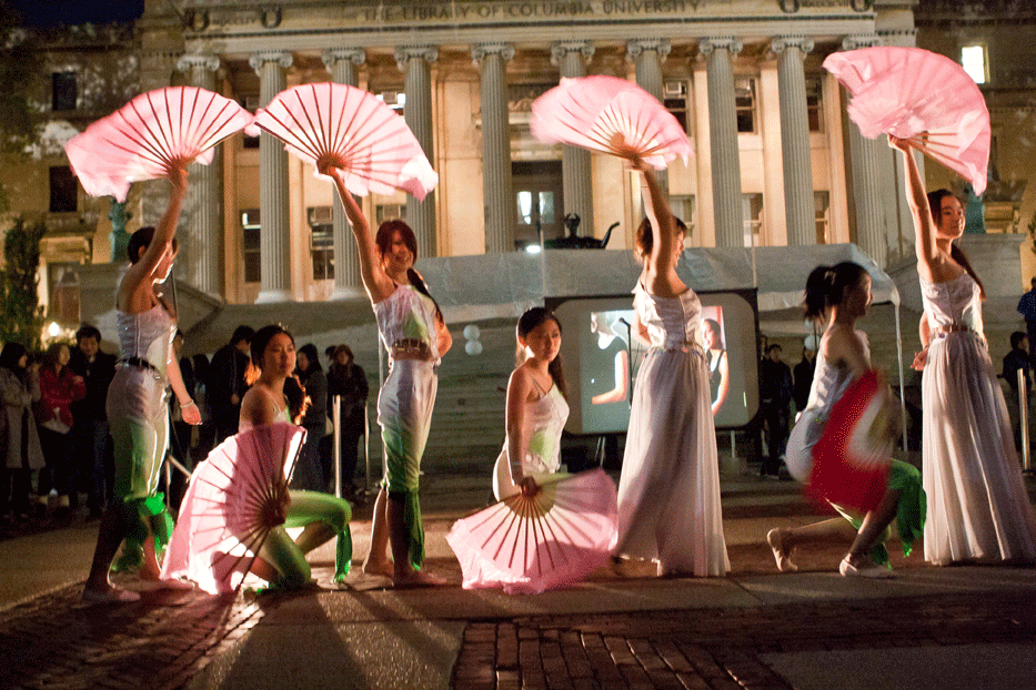 A group of young women perform a fan dance on the plaza at night.