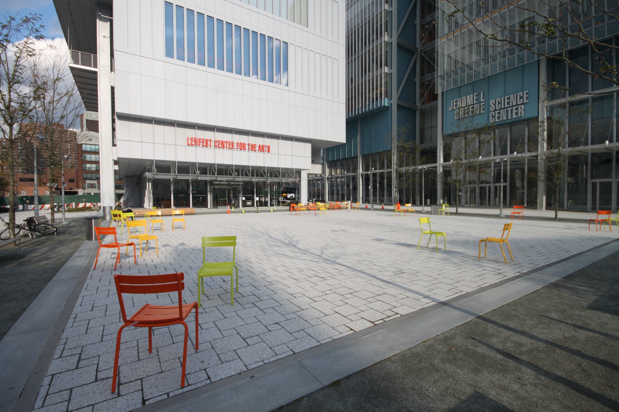 A grey stone plaza outside Lenfest Center for the Arts, a concrete structure, and Jerome L. Greene Science Center, a glass and steel structure. Multi-color metal chairs dot the plaza.