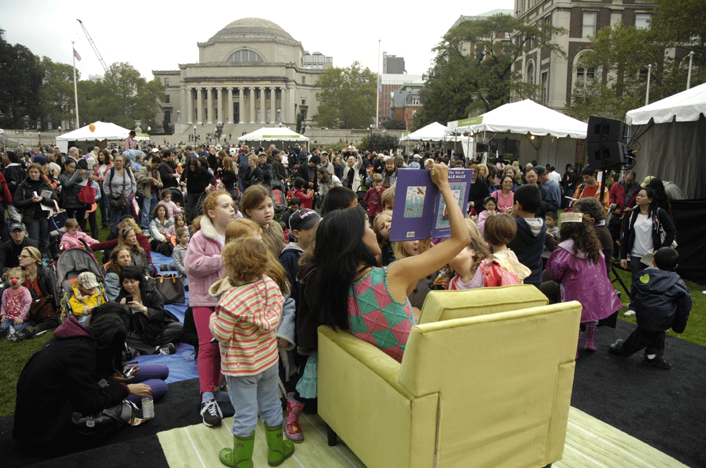 A woman sitting in a yellow arm chair on a raised platform holds up a picture book to a crowd of parents and their young children. In the background, a grand columned building can be seen.