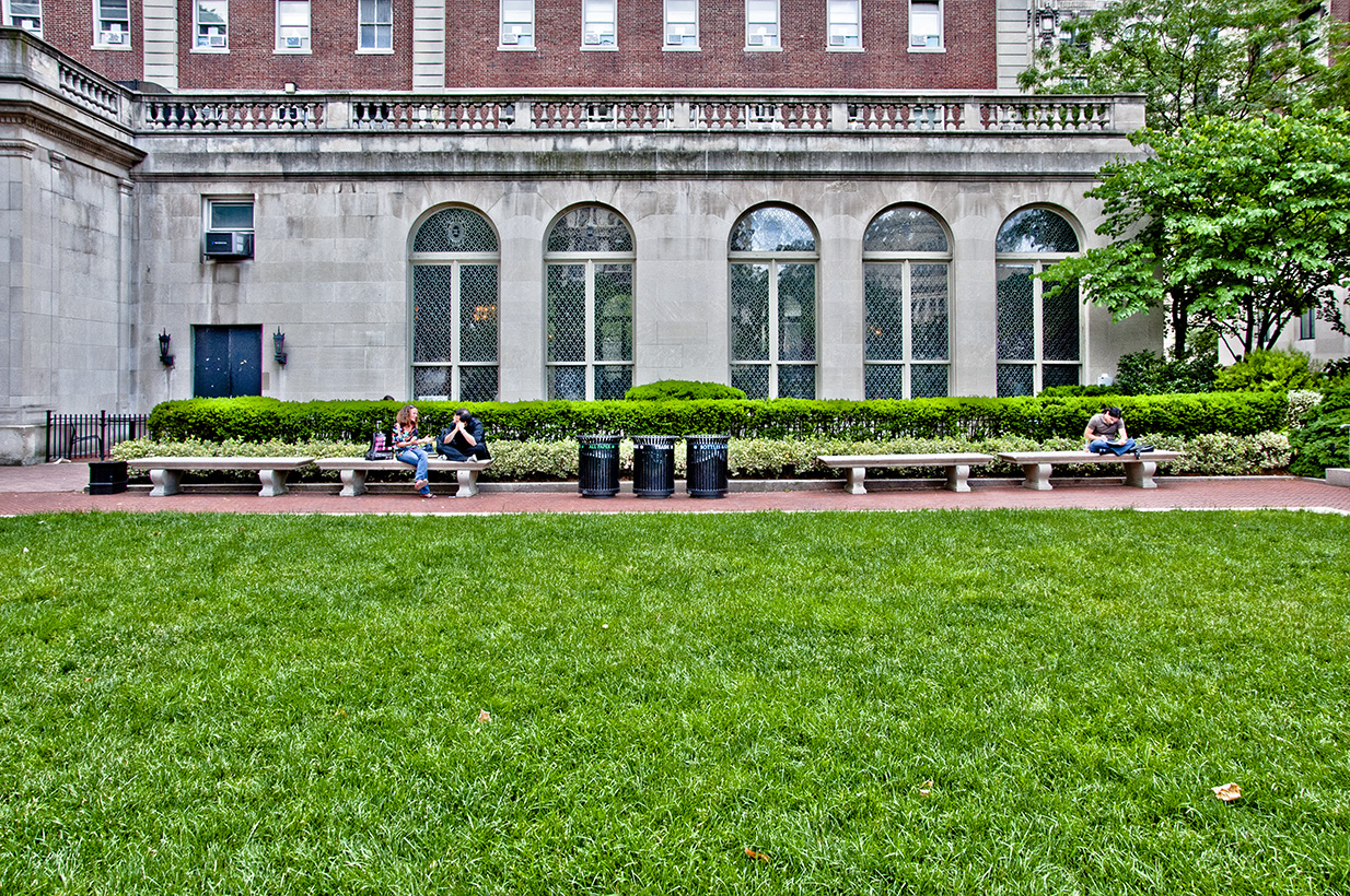 A small lawn with near a brick and stone building. People sit on benches at the outskirt of the lawn.