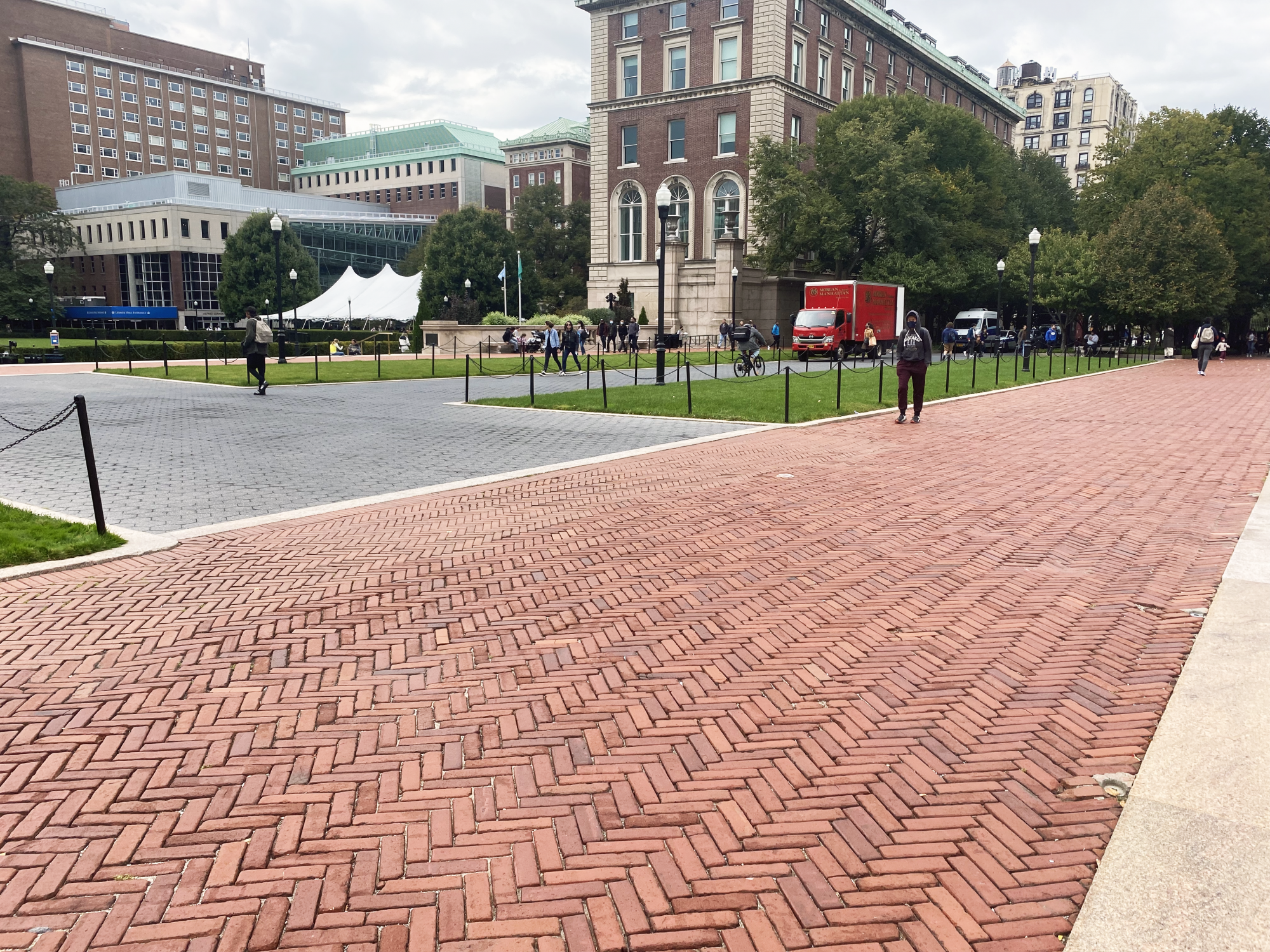 A red brick pathway with green lawns offsetting a gray cobblestone center. Trees and buildings in the background and people walking in the foreground.