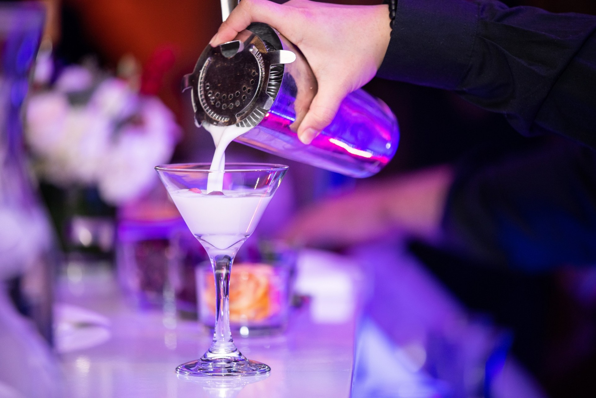 A bartender pours a white drink from a cocktail shaker into a martini glass