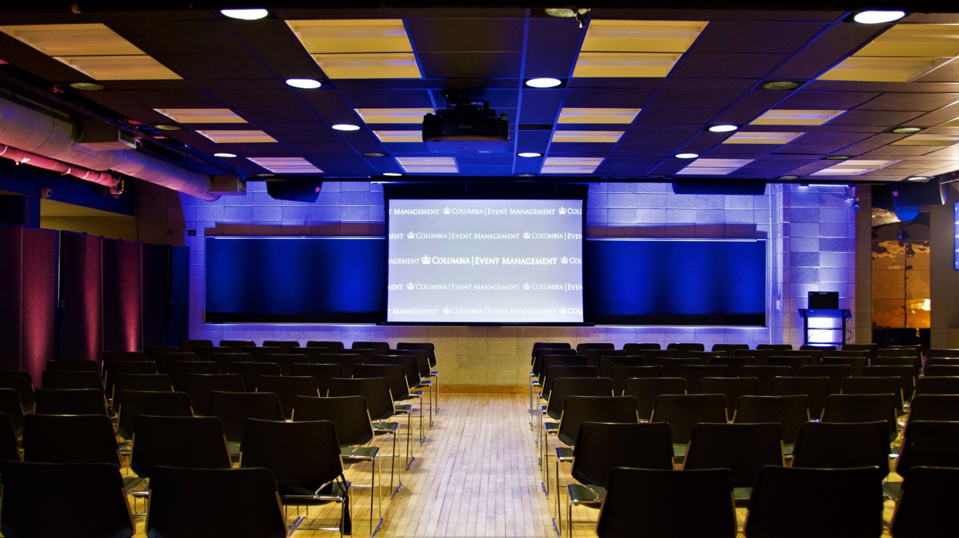 Rows of black chairs face a projector screen mounted on a stone wall.
