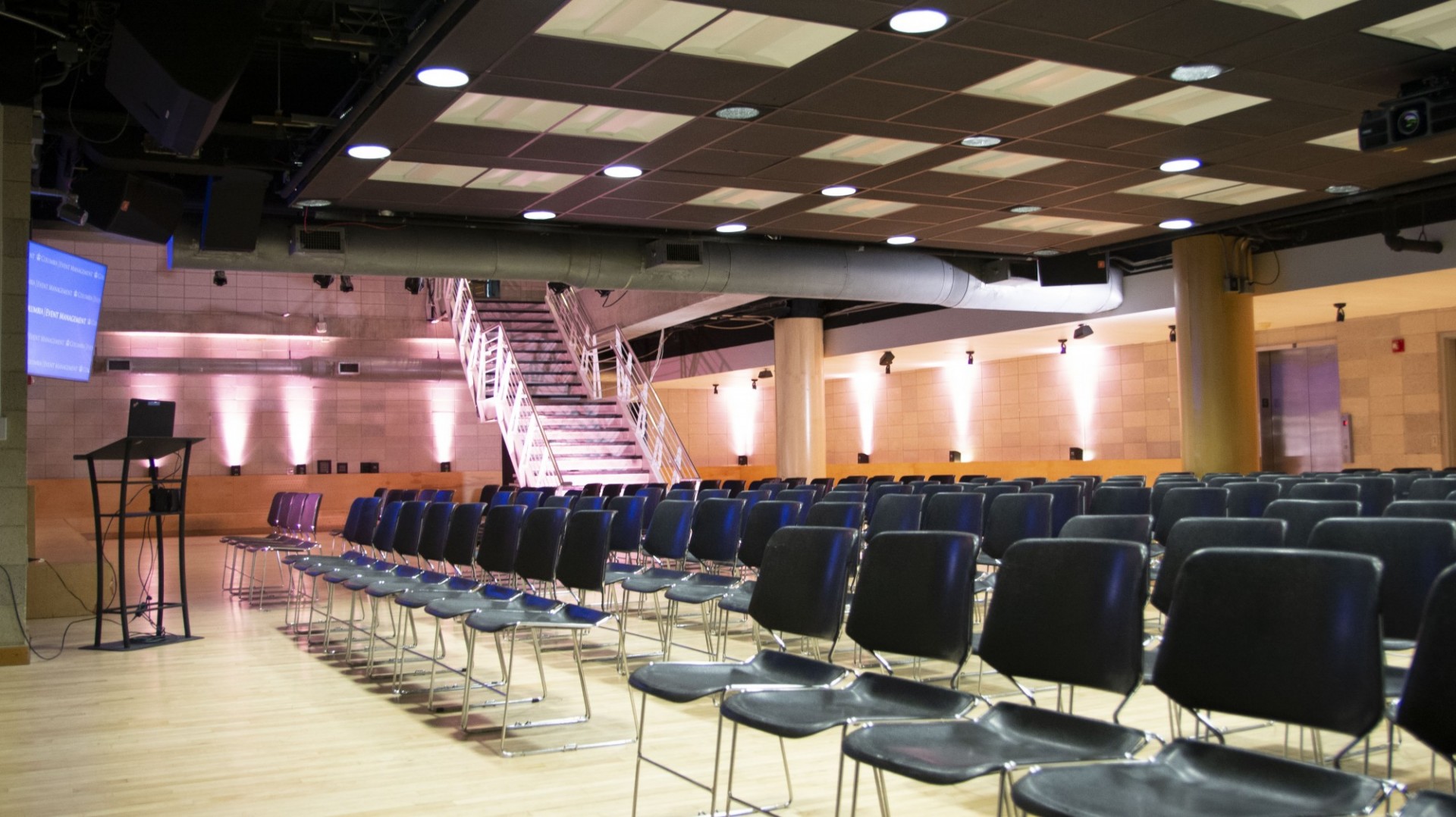 Rows of black chairs face a podium. In the background, a staircase can be seen.