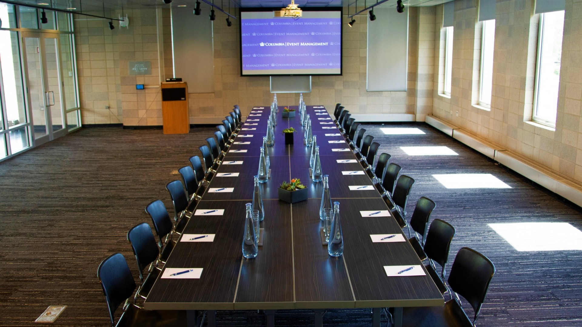 A long, rectangular conference table is in the middle of a grey-carpeted room. The table is set with water glasses and pitchers. A projector screen is mounted on a wall perpendicular from the table.