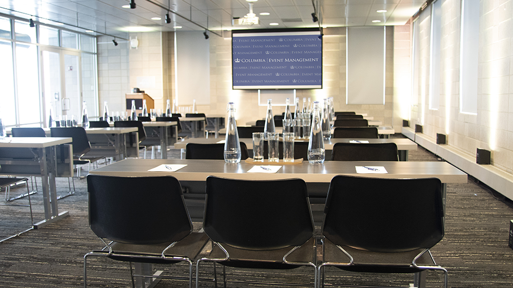 Rows or tables set with three chairs and water glasses and pitchers face a projector screen and podium.