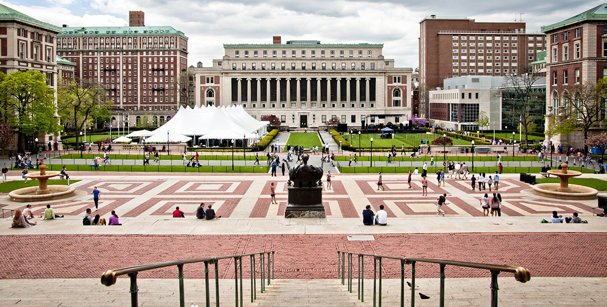 A southern-looking view of Low Plaza taken from Alma Mater during the day. In the background, campus is bustling. People are walking across the plaza and College Walkway, and tents have been erected on a southern lawn.