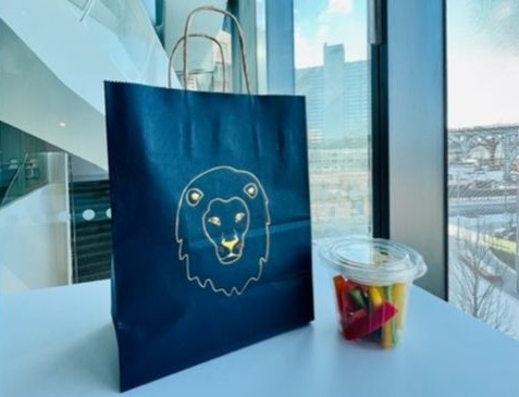 A blue paper bag with an embossed gold lion outline sits next to a cup of assorted vegetable sticks in front of a window overlooking Manhattanville Campus