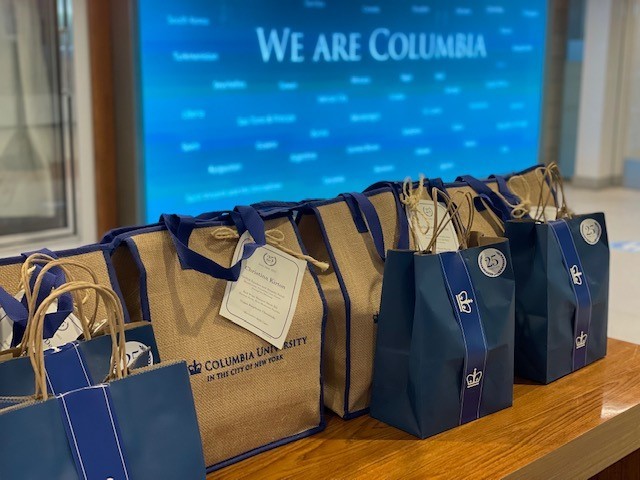 Reusable tan tote bags and navy blue paper bags holding boxed dinners for the Columbia University 25 Year Club celebration sit on a table ready for pickup.
