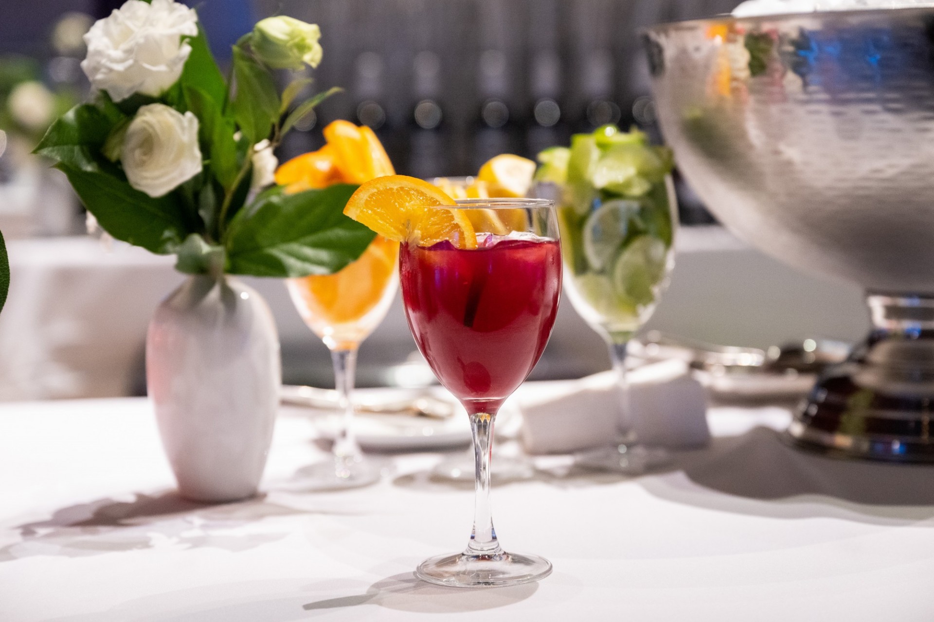 A red cocktail with an orange slice on the rim sits on a white table with a bouquet of white flowers in the background