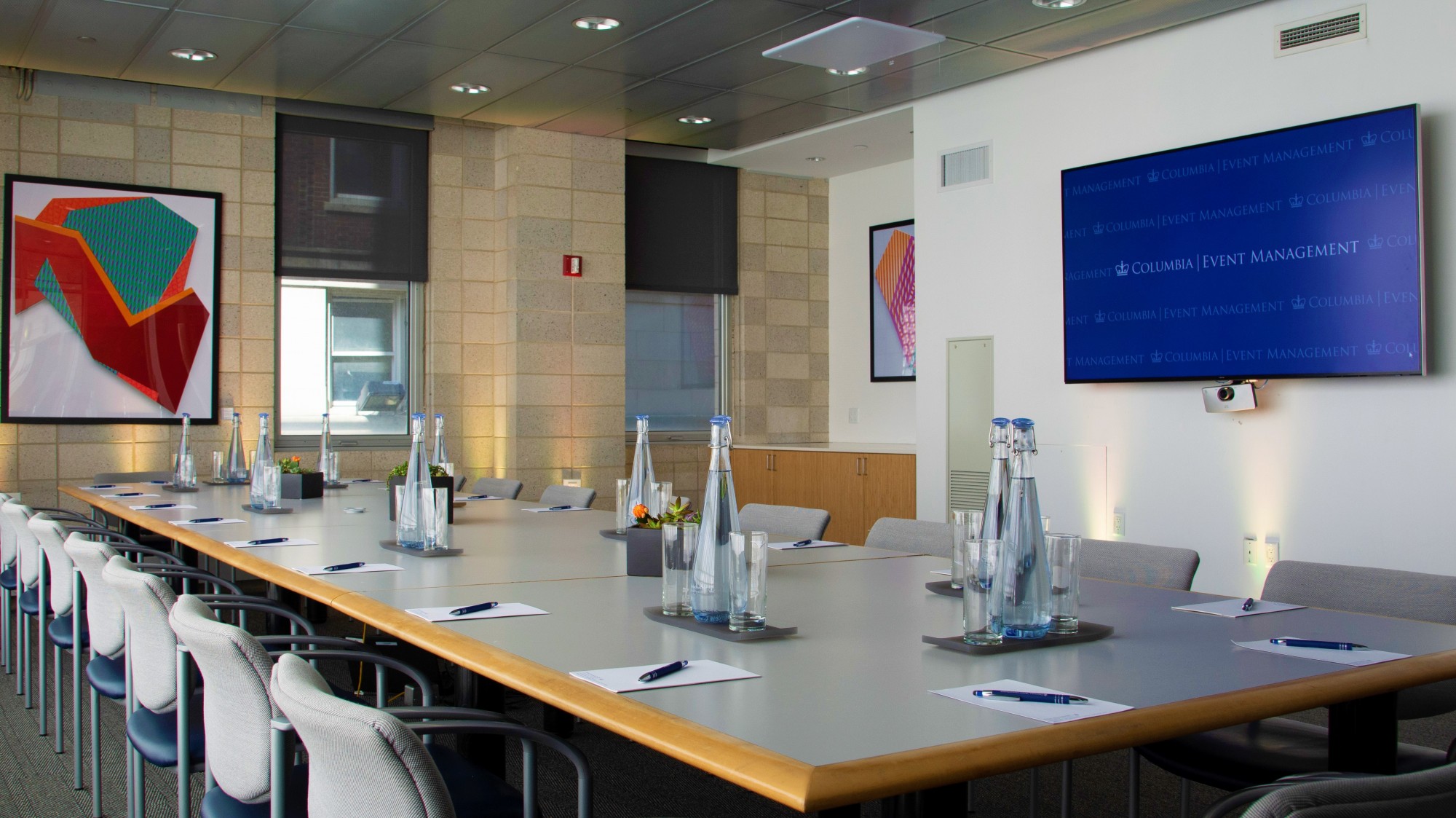 A long, rectangular grey conference table bisects the room. The table is set with glasses and water pitchers. A TV screen is mounted on a white wall perpendicular to the table.