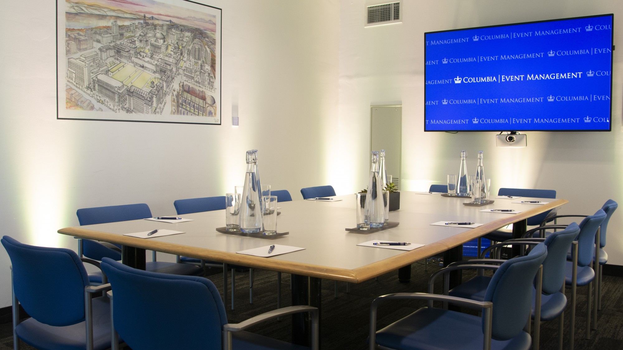 A grey, rectangular table sits in the middle of a white walled room. The table is set with glasses and water pitches and 12 blue chairs. A TV screen is mounted on the wall and faces the table.
