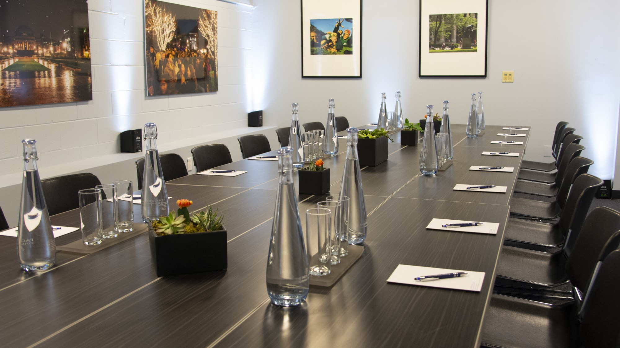 A dark, rectangular conference table sits in the middle of white walled room. The table is set with water glasses and pitchers and decorative plants. Artwork adorns the walls.
