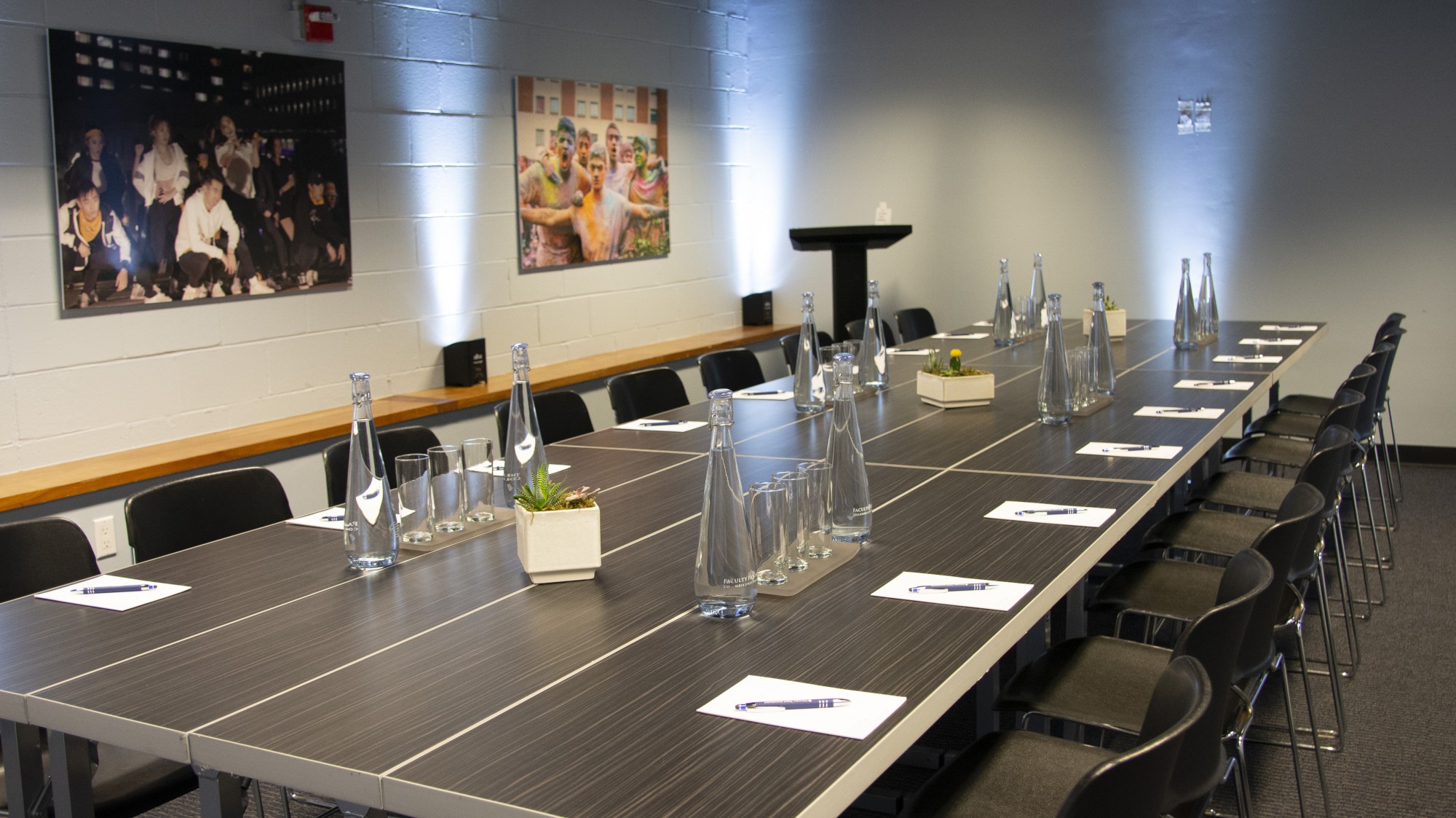 A dark, rectangular conference table sits in the middle of white walled room. The table is set with water glasses and pitchers and decorative plants. Artwork adorns the walls. A podium is stationed in one corner of the room.
