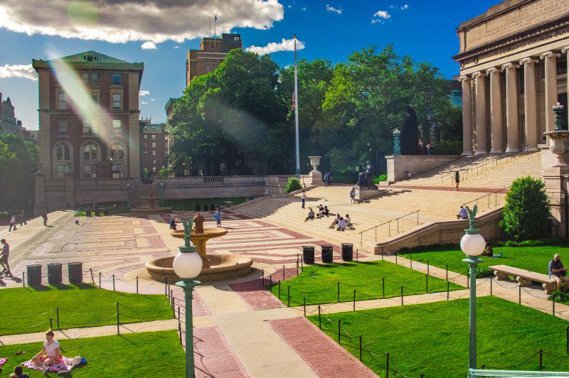 A grey and red brick patterned plaza sits in front of grand stairs and an ornate grey stone, columned building. People are walking across the plaza and sitting on the stairs.