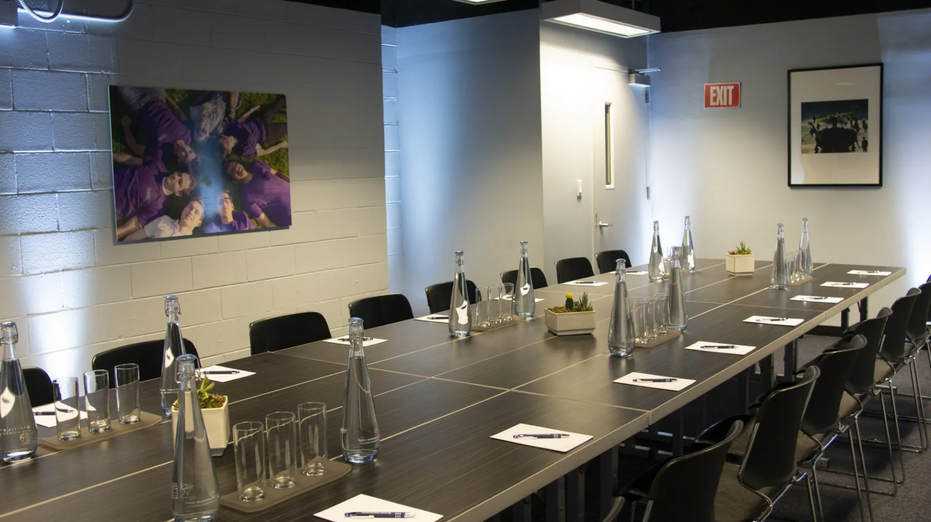 A dark, rectangular conference table sits in the middle of white walled room. The table is set with water glasses and pitchers and decorative plants. Artwork adorns the walls.