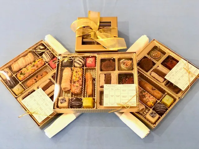 Gold partitioned boxes contain Foie Gras Eclair, Mini Lobster Roll, Mushroom Beignet, Cured Salmon Cucumber Roll, English Tea Pinwheel, Stilton Beef Rosette, Tamari Glazed Peanut Crumb Tofu, Watermelon Ahi Tuna, and Assorted Petit Fours.