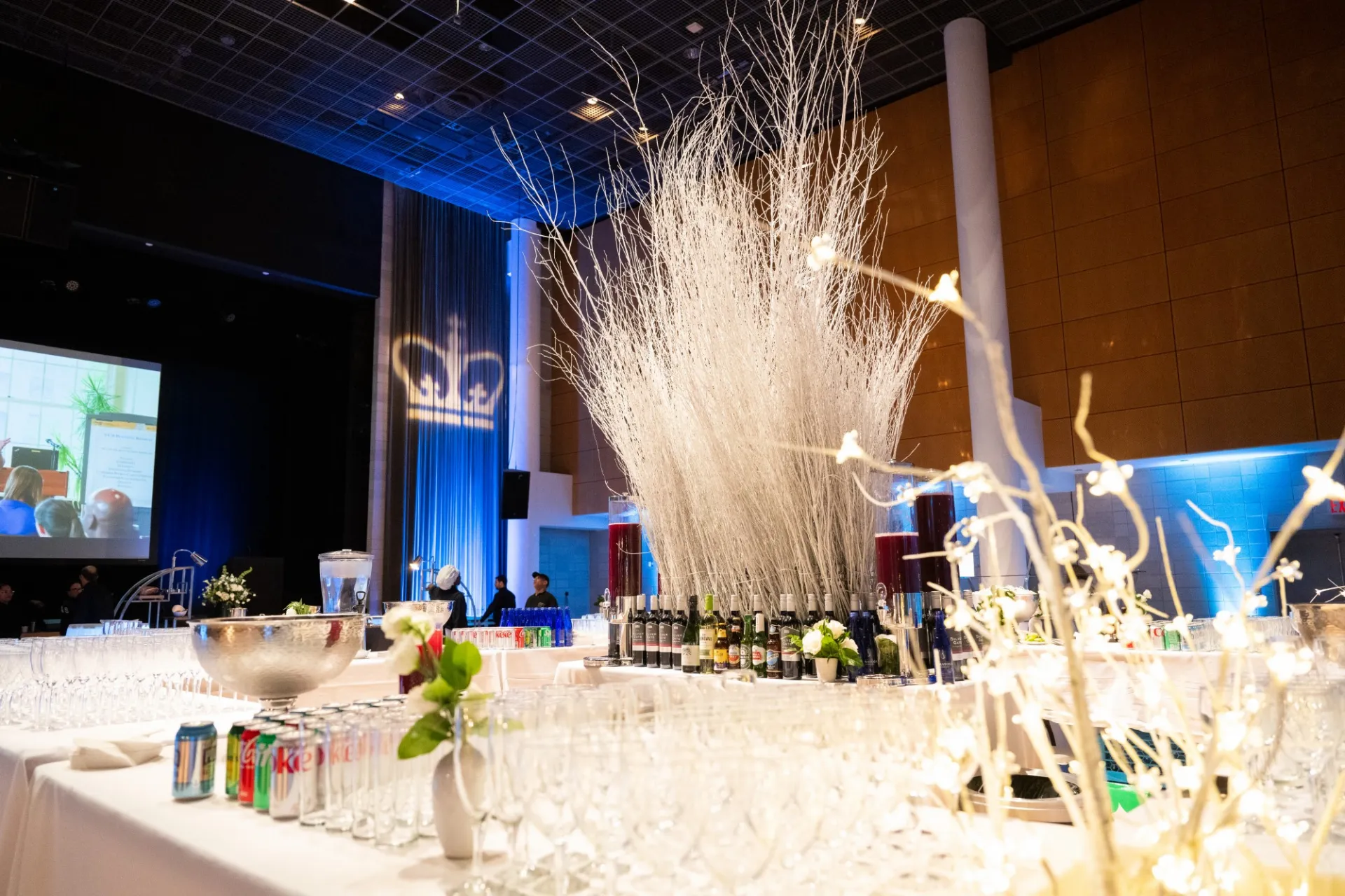 A table clad in white linen displays a beverage station in Roone Arledge Auditorium, with festive holiday lighting and blue curtains in the background