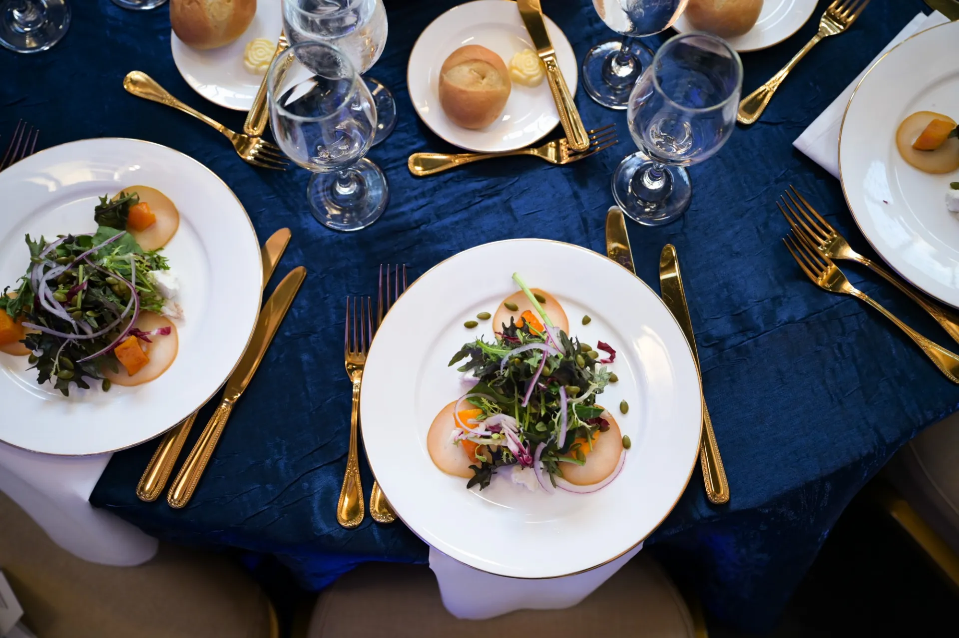 A rich blue tablecloth, gold cutlery, and plated first course salads await guests at an elegant event
