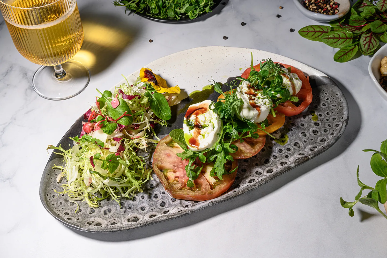 tomato and burrata salad with frisee and a glass of wine on a white tablecloth surrounded by herbs and spices