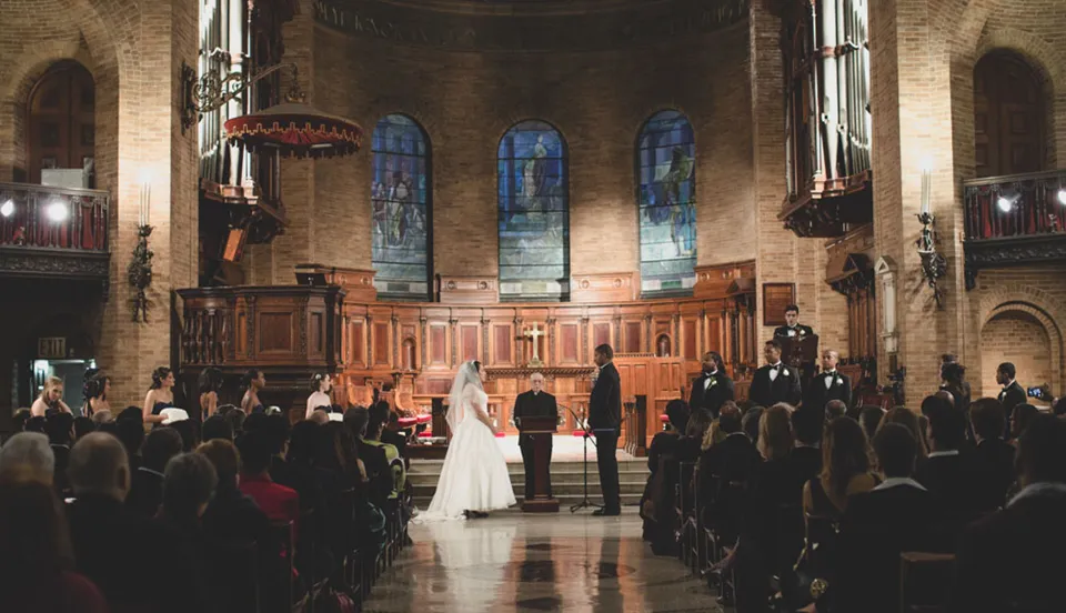 A wedding ceremony takes place inside Saint Paul's Chapel. A bride and groom stand opposite each other with the officiant in the middle, facing the camera. Guests are seated in rows along each side of the aisle. Stained glass windows adorn the back wall of the chapel.